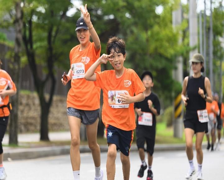 주황색 글로벌 6K 티셔츠를 입은 아이와 어른 참가자들이 도로 위를 함께 달리며 손짓하고 있다.