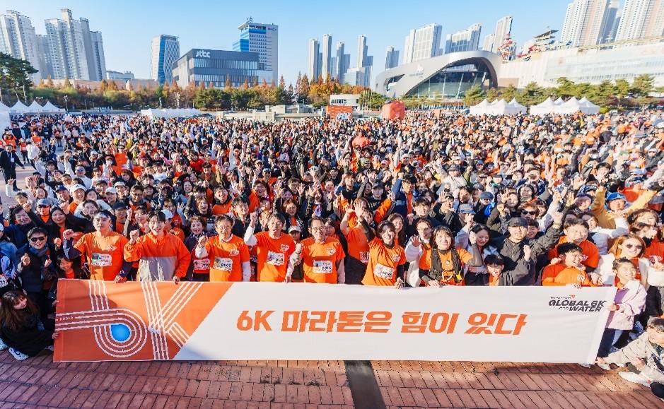 대규모 참가자들이 모인 글로벌 6K 행사 현장에서 ‘6K 마라톤은 힘이 있다’라는 현수막 앞에 서 있다.